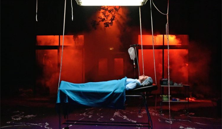 Une jeune femme est allongée sur un lit d'urgence psychiatrique, sous une lumière froide, alors que tout autour d'elle donne une ambiance de fête.